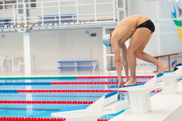 Young muscular swimmer in low position on starting block in a swimming pool — Stock Photo, Image