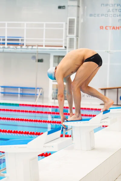 Young muscular swimmer in low position on starting block in a swimming pool — Stock Photo, Image