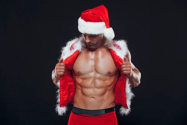 Pai Natal sexy. Fisiculturista jovem bonito santa cláusula sorriso vestindo chapéu de Papai Noel mostra cubos de abdominais na véspera de Ano Novo e Natal férias de inverno fundo preto. — Fotografia de Stock