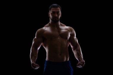 Siyah arka planda, kaslarını göstermek için yumruklarını sıkmış bir gölgede duran bir vücut geliştiricinin portresi.