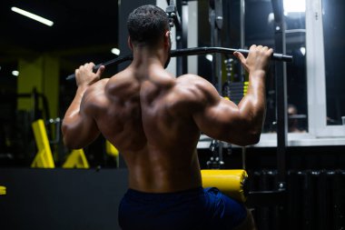 Yakışıklı fitness adamı spor salonunda, arka planda üst blok makinesini kullanarak egzersiz yapıyor.