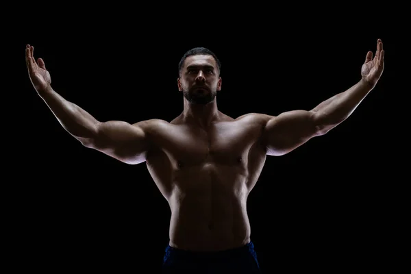 Portrait d'un bodybuilder debout isolé sur fond noir dans une ombre avec les mains levées pour montrer ses muscles — Photo