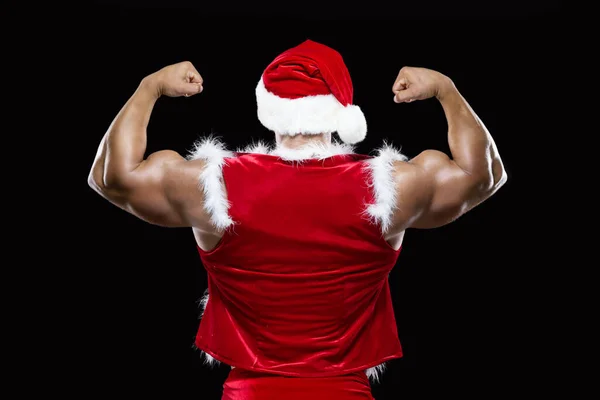 Widok z tyłu młody Święty Mikołaj w czerwonym kapeluszu Bożego Narodzenia pokazując swoje silne bicepsy izolowane na czarnym tle — Zdjęcie stockowe
