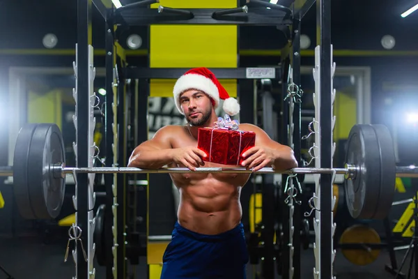 Молодий привабливий Санта Клаус у Різдвяному капелюсі тримає подарунок на дзвіночку в спортзалі, спереду. — стокове фото