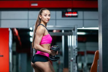 Modern spor salonunda kollarını esneten halterlerle ağırlık kaldıran güçlü, kaslı genç bir kadının yan görünüşü.