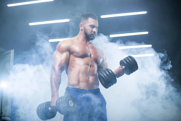 Front view of a muscular bodybuilder lifting dumbbells in blue shorts posing in a gym standing in smoke — Stock Photo, Image