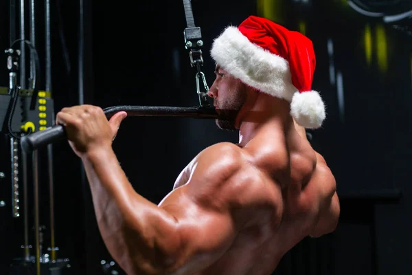 Красивий фітнесмен у різдвяній шапці виконує зворотні тренування, використовуючи тягу верхньої блокової машини в спортзалі, вид ззаду — стокове фото