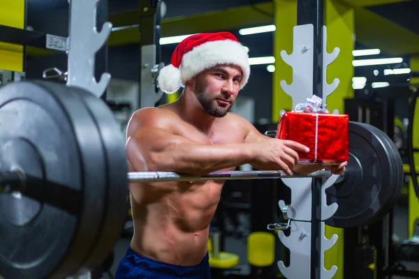 Молодий привабливий Санта Клаус у Різдвяному капелюсі тримає подарунок, спираючись на дзвіночок у спортзалі. — стокове фото