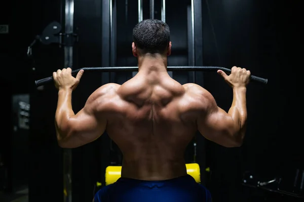 Bel homme de remise en forme effectue des séances d'entraînement arrière en utilisant la poussée de la machine bloc supérieur dans une salle de gym, vue arrière — Photo
