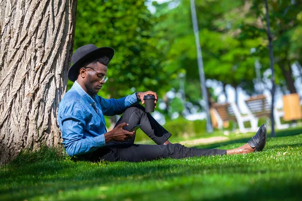 Şapkalı ve gözlüklü genç bir adamın portresi parktaki ağacın altında oturuyor. Bir elinde kahve, diğer elinde cep telefonu var. — Stok fotoğraf