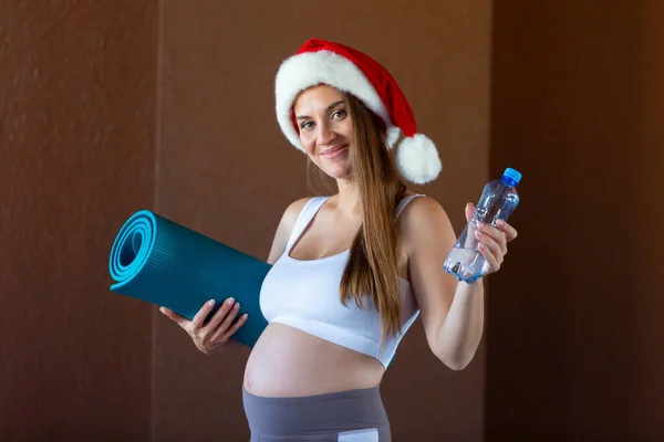 Улыбающаяся беременная женщина в рождественской шляпе держит бутылку с водой и коврик, готовый к упражнениям — стоковое фото