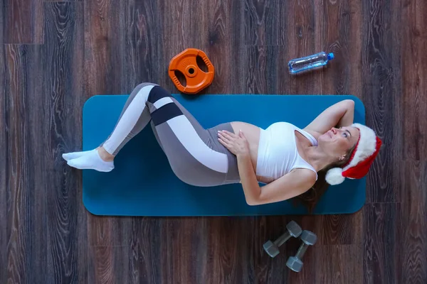 Κάτοψη της νεαρής όμορφης εγκύου στο καπέλο του Άη Βασίλη ξαπλωμένη στο χαλάκι στο πάτωμα σε ένα γυμναστήριο κοντά σε αλτήρες, ένα μπουκάλι νερό και ένα δίσκο — Φωτογραφία Αρχείου