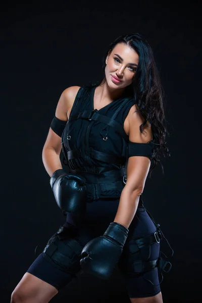 Een jonge atletische vrouw in een EMS pak met bokshandschoenen op een geïsoleerde zwarte achtergrond. EMS-opleiding. Elektro gespierde stimulatie machine. — Stockfoto