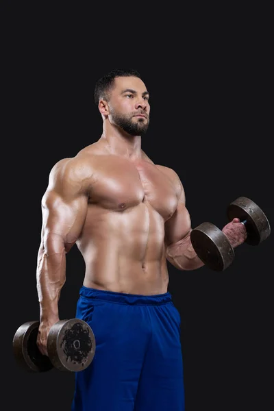 Young muscular sportsman is lifting heavy dumbbells showing his strong biceps isolated on black backgrounds — Stock Photo, Image