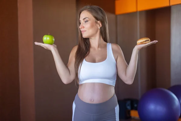 Joven chica atractiva está haciendo la elección entre comida sana y dañina sosteniendo una manzana verde y una hamburguesa en sus manos. Concepto de nutrición adecuada, comida saludable, comida rápida —  Fotos de Stock