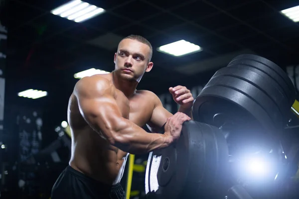 Joven atleta muscular hombre de apariencia modelo está descansando después de ejercitar los músculos del entrenamiento en el gimnasio de pie junto a los discos —  Fotos de Stock