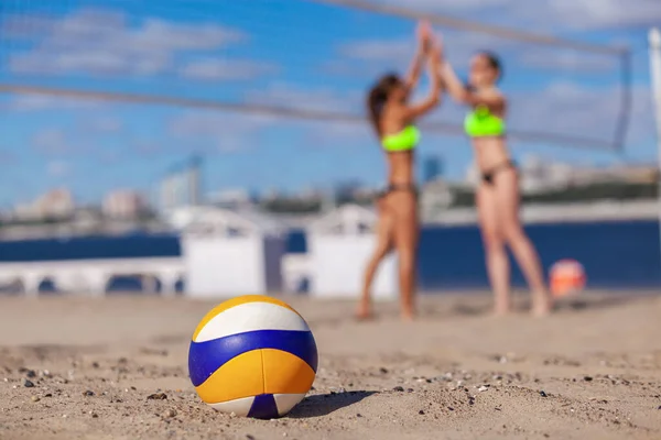Close up de uma bola na areia em um fundo embaçado de duas meninas felizes desfrutando de uma vitória no vôlei de praia — Fotografia de Stock