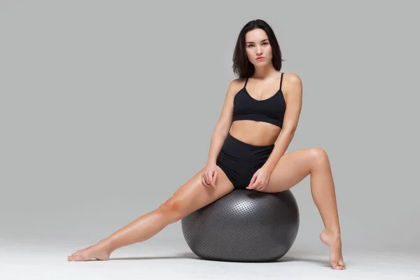 Portret pełnej długości sportowej seksownej dziewczyny siedzącej na piłce fitness rozciągającej swoją nogę na szarym tle — Zdjęcie stockowe