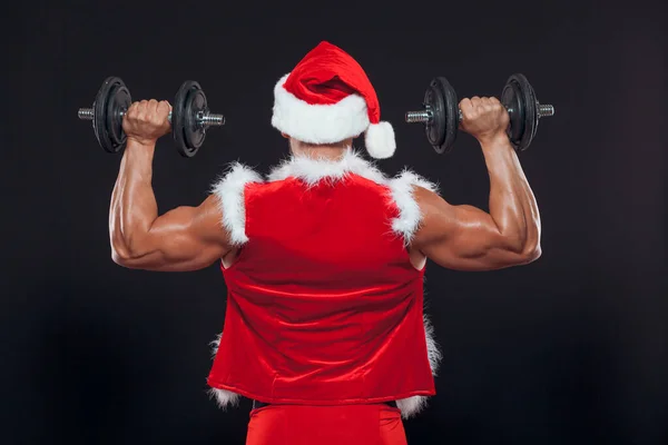 Sexy Santa Claus. Bodybuilder young handsome santa clause smile holds a dumbbells at New Years eve and Christmas winter holiday black background. The view from the back.