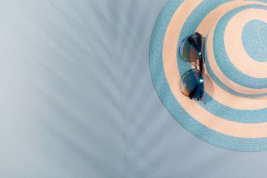 Turkuaz mavi arka planda plaj aksesuarları. Güneş gözlükleri ve palmiye yaprağının gölgesinde çizgili mavi şapka. Plaj tatili kavramı, deniz turu, sıcak güneşli yaz. Reklam alanı