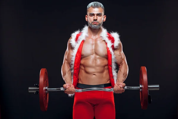 Sexy Santa Claus. Bodybuilder young handsome santa clause smile train muscles arms and biceps at New Years eve and Christmas winter holiday black background.