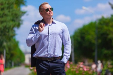 Mavi takım elbiseli genç yakışıklı adam güneş gözlükleriyle parkta yürüyor. Ceketini omuzunda tutuyor.