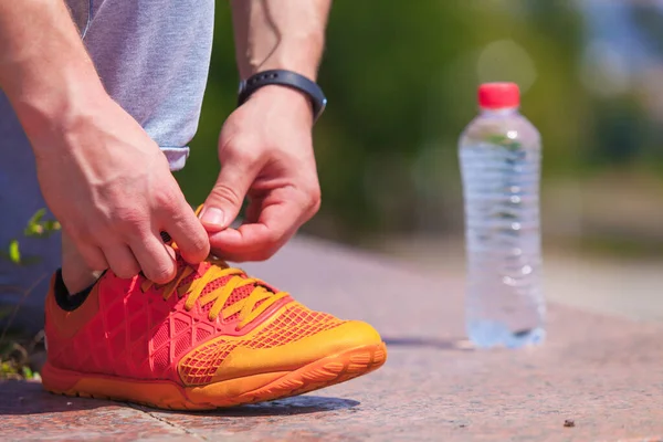 Close up on mans hands tying shoelaces on orange sneakers and a bottle of water during a run in a summer park — Stock Photo, Image