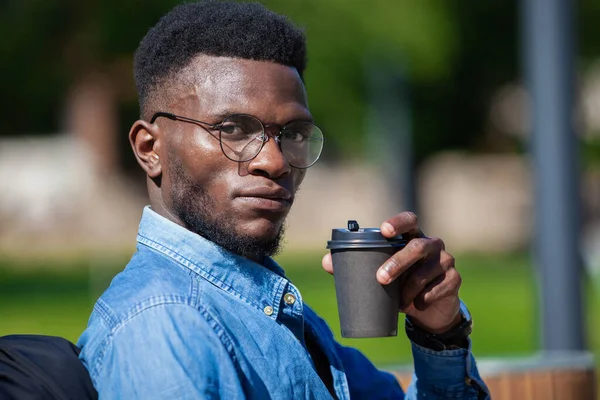 Közel a fiatal afro-amerikai üzletember elegáns ruhákat ül padon egy városi parkban, és iszik forró kávét — Stock Fotó