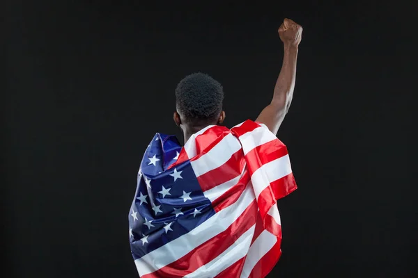 Visão traseira do homem vestindo bandeira americana de pé com a mão levantada isolada sobre fundo preto. Conceito de patriotismo, amor pela Pátria, símbolo nacional — Fotografia de Stock