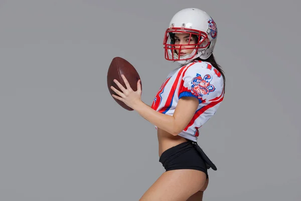 Sporty girl wearing sexy uniform of American football player and helmet posing with ball ready to throw isolated on grey background