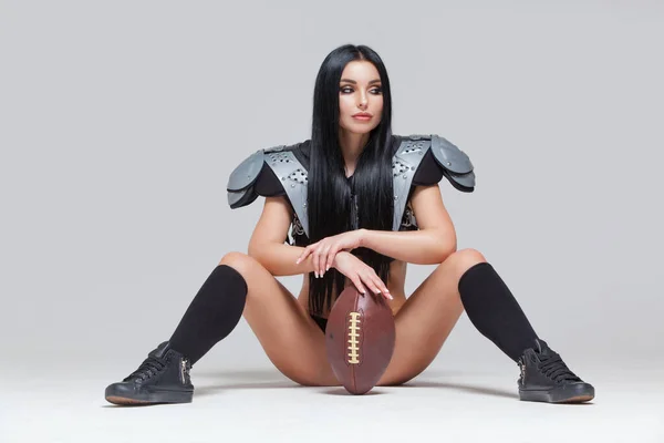 Hermosa morena deportiva vestida con bikini y uniformes de fútbol americano posando sentada en el suelo con una pelota entre las piernas aislada sobre fondo gris — Foto de Stock
