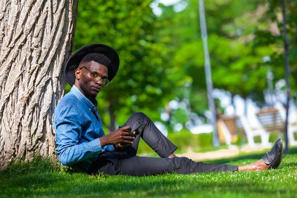 Şapkalı ve gözlüklü genç bir adamın portresi parktaki ağacın altında oturuyor bir elinde kahve, diğer elinde cep telefonu var. — Stok fotoğraf