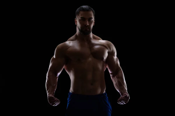 Portret van een bodybuilder geïsoleerd op zwarte achtergrond in een schaduw met gebalde vuisten om met zijn spieren te pronken — Stockfoto