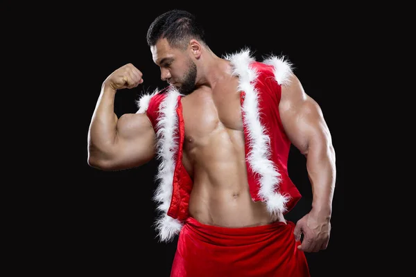 Jeune Père Noël sexy dans un costume de carnaval rouge sur un corps nu montre ses forts biceps isolés sur fond noir — Photo