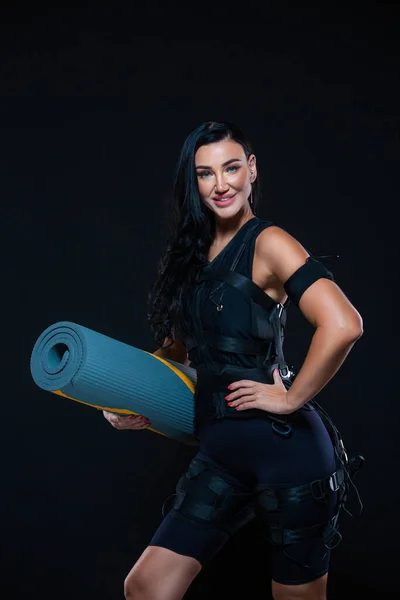 Uma jovem atlética num fato EMS com um tapete num fundo preto isolado. Treinamento EMS. Máquina de estimulação eletromuscular. — Fotografia de Stock