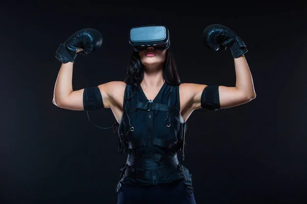 A young athletic woman in an EMS suit and virtual reality glasses with Boxing gloves on an isolated black background. EMS training. Electrical muscle stimulator.