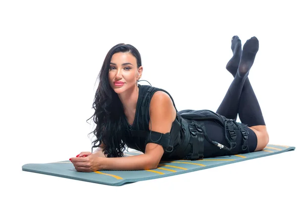 En ung atletisk kvinna i en EMS kostym ligger på en matta på en isolerad vit bakgrund. Medicinsk utbildning. Elektrisk muskelstimulator. — Stockfoto