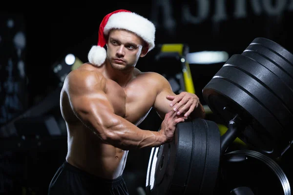 Молодий м'язовий Санта Клаус у різдвяному капелюсі відпочиває після роботи над тренувальними м'язами в спортзалі, що стоїть біля дисків. — стокове фото