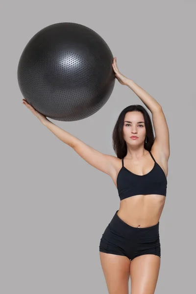 Portret kobiety w czarnej odzieży sportowej trzymającej piłkę treningową nad głową na szarym tle — Zdjęcie stockowe