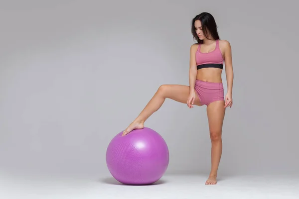 Jong aantrekkelijk meisje in moderne roze sportkleding poseert zetten haar voet op fitness bal geïsoleerd op grijze achtergrond. Totale lengte — Stockfoto