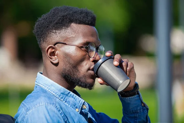 Közel a fiatal afro-amerikai üzletember elegáns ruhákat ül padon egy városi parkban, és iszik forró kávét — Stock Fotó