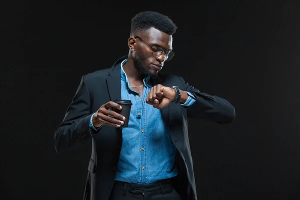 Портрет американского африканского бизнесмена, держащего чашку кофе и смотрящего на часы, изолированные на черном фоне — стоковое фото