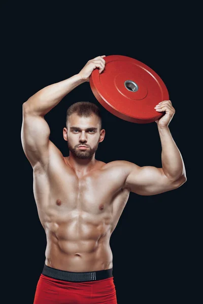 Portrait of sexy muscular male with a barbell disc raised above his head isolated on black background — Stock Photo, Image