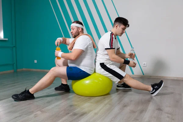 胖男人和他的私人教练在健身班的艰苦训练后，坐在健身球上，拿着瓶装水。那个超重的男人非常疲倦 — 图库照片