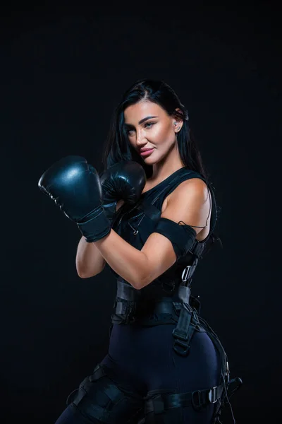 En ung atletisk kvinna i en EMS kostym med Boxningshandskar på en isolerad svart bakgrund. Medicinsk utbildning. Elektromuskulär stimuleringsmaskin. — Stockfoto