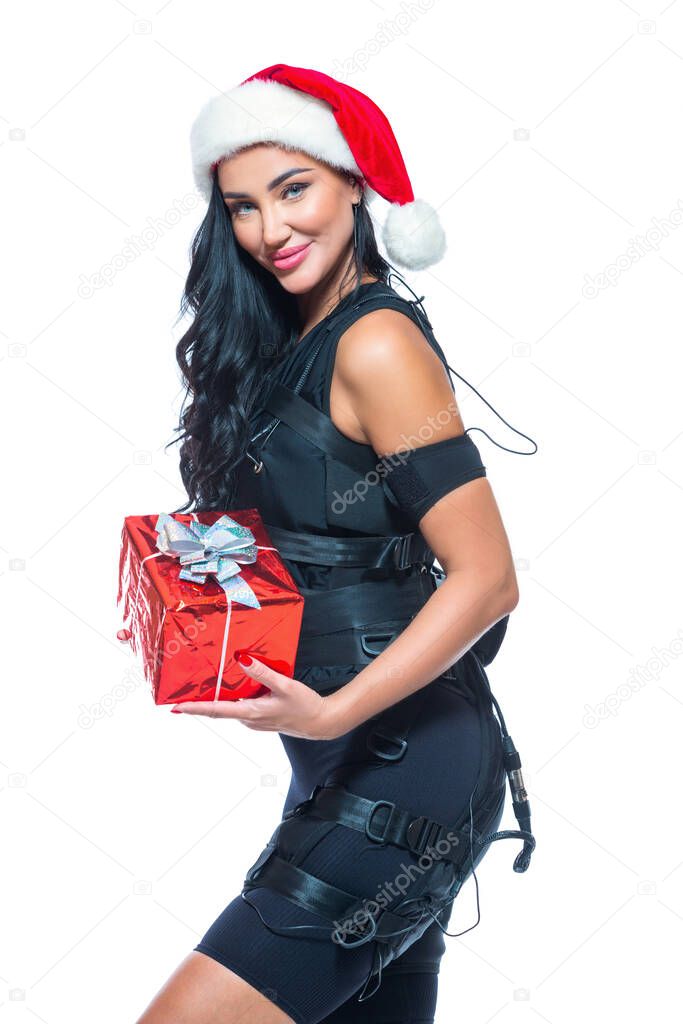 Christmas. A young athletic woman in an EMS suit and Santa Claus hat holds a gift box on an isolated white background. EMS training. Electrical muscle stimulator.