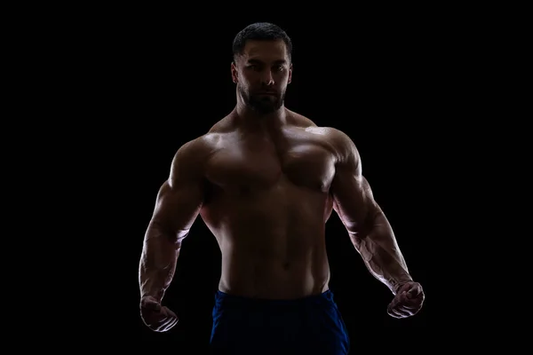 Porträt eines Bodybuilders, der isoliert auf schwarzem Hintergrund im Schatten steht und seine Muskeln mit geballten Fäusten zur Schau stellt — Stockfoto