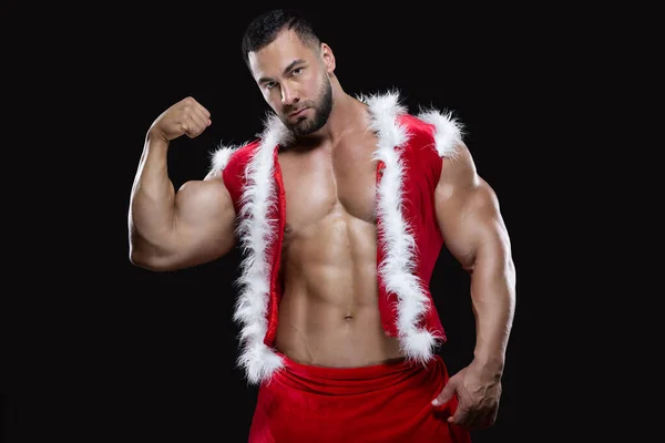 Joven Santa Claus sexy en un disfraz de carnaval rojo en un cuerpo desnudo muestra sus fuertes bíceps aislados sobre fondo negro — Foto de Stock
