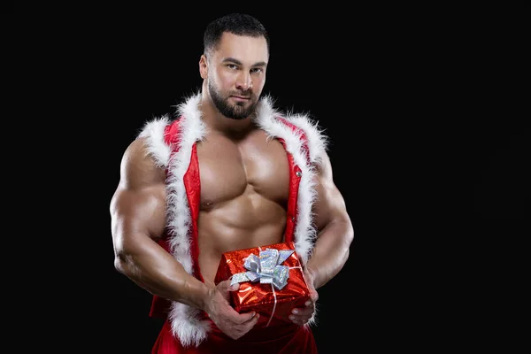 Muscular sexy sportsman in a red Santa Claus costume over naked body is holding a Christmas gift isolated on black background — Stock Photo, Image