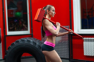 Modern bir spor salonunda omzunda balyozla lastiğin yanında poz veren kaslı bir kadın.